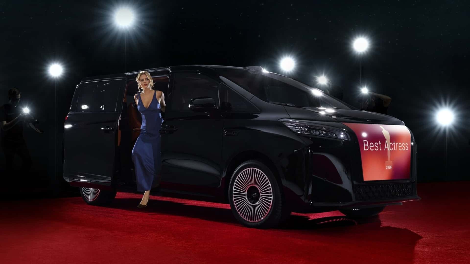 Kvinde i blå kjole stiger ud af elektrisk bil på den røde løber omgivet af fotografer