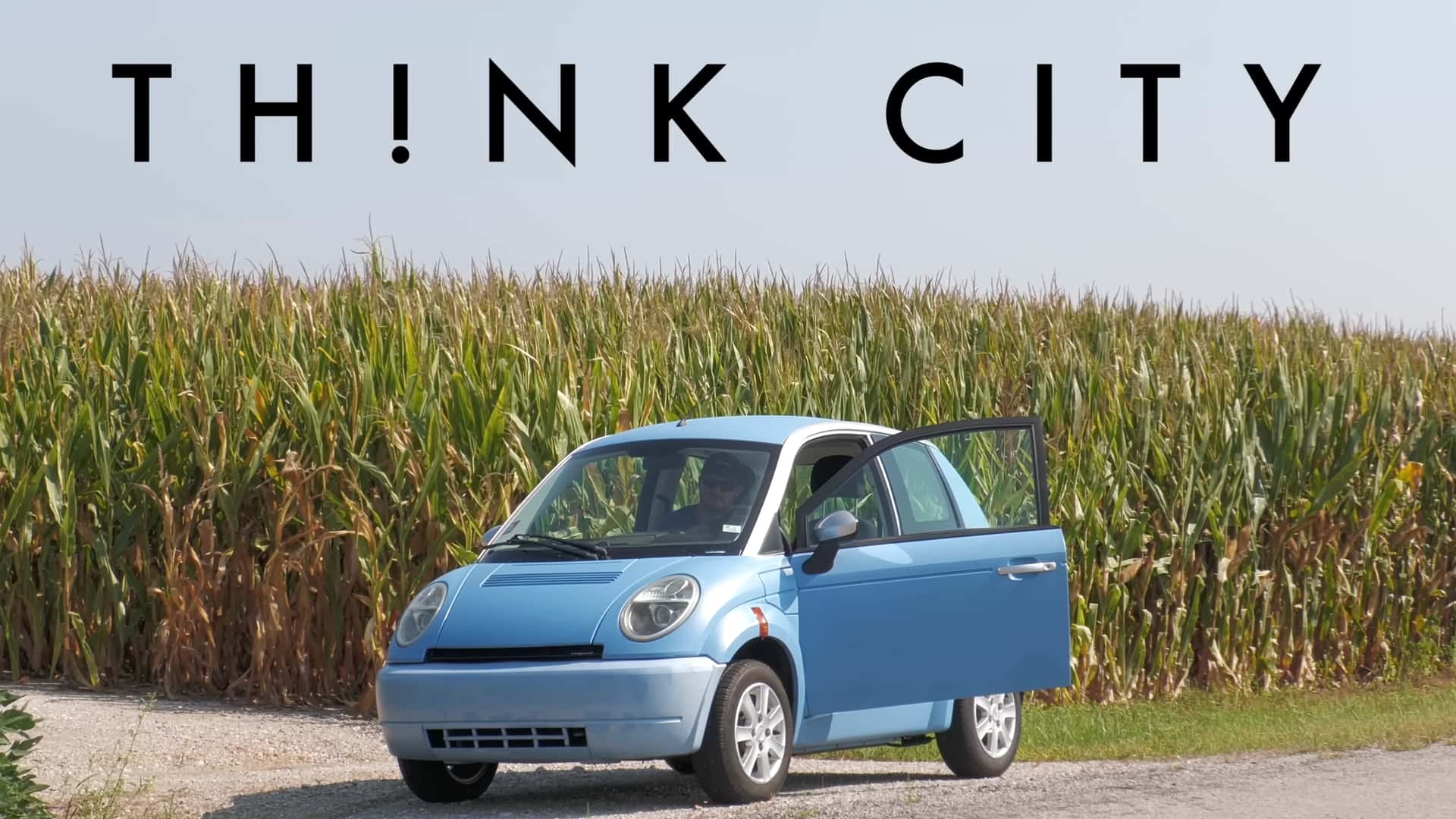 Blå Think City elbil parkeret foran en majsmark