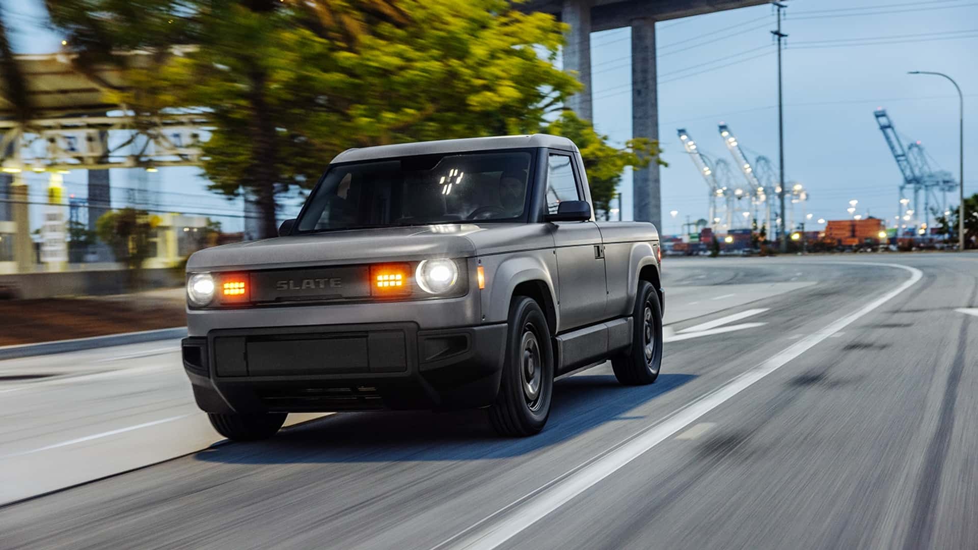 Elektrisk pickup kører langs en byvej med havnekraner i baggrunden