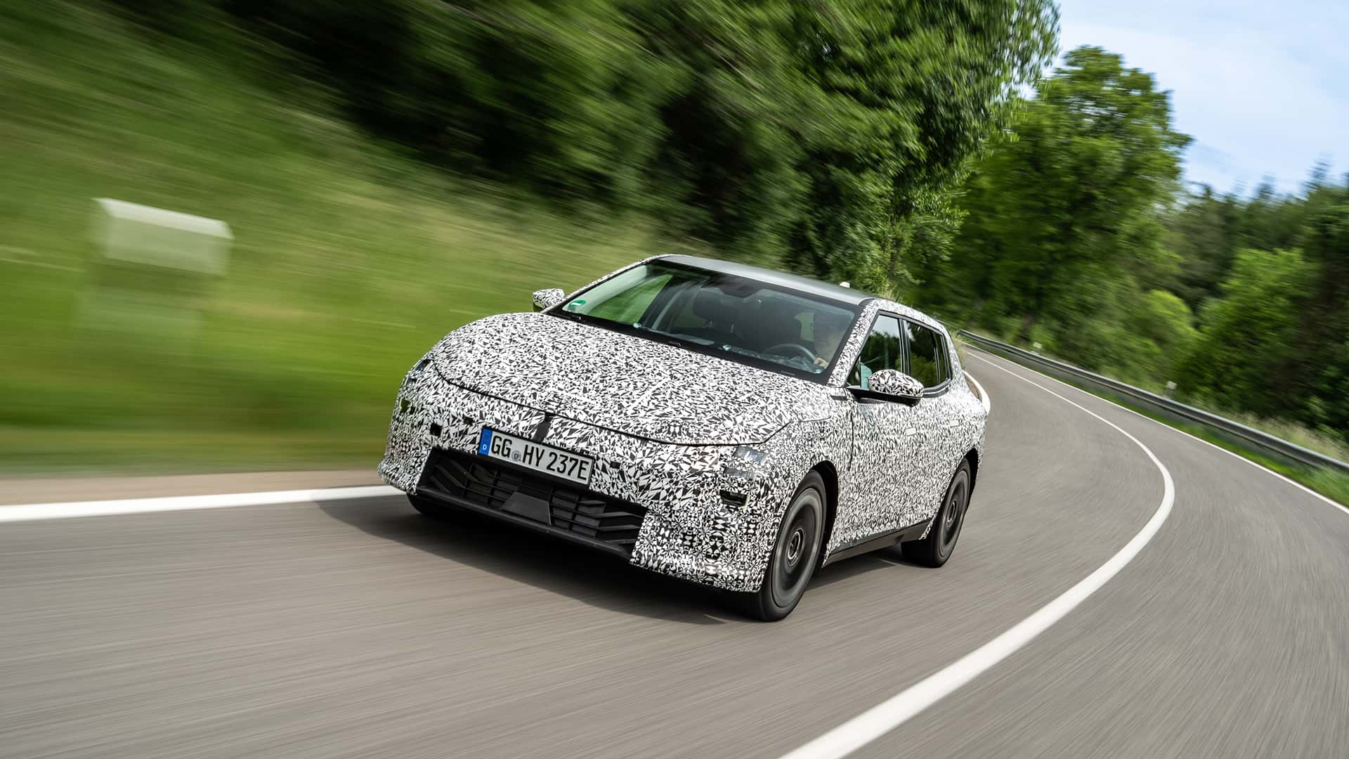 Camufleret elbil kører på en snoet vej omgivet af grønne træer