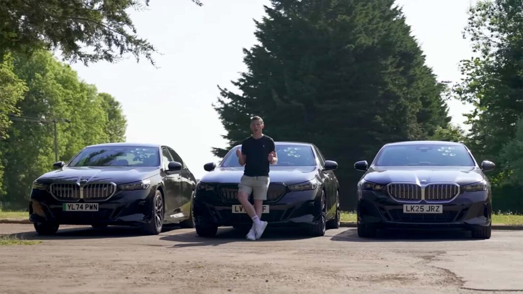 Tre sorte biler parkeret side om side med en person stående ved den midterste bil, omgivet af træer i baggrunden.