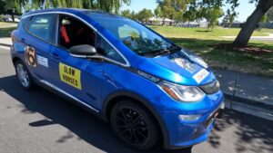 Blå Chevrolet Bolt EV med klistermærker på siden, parkeret i solrigt område