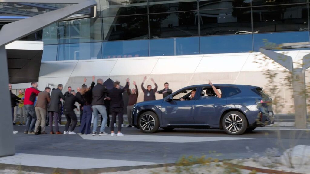 Gruppe mennesker fejrer foran en elektrisk bil ved moderne bygning