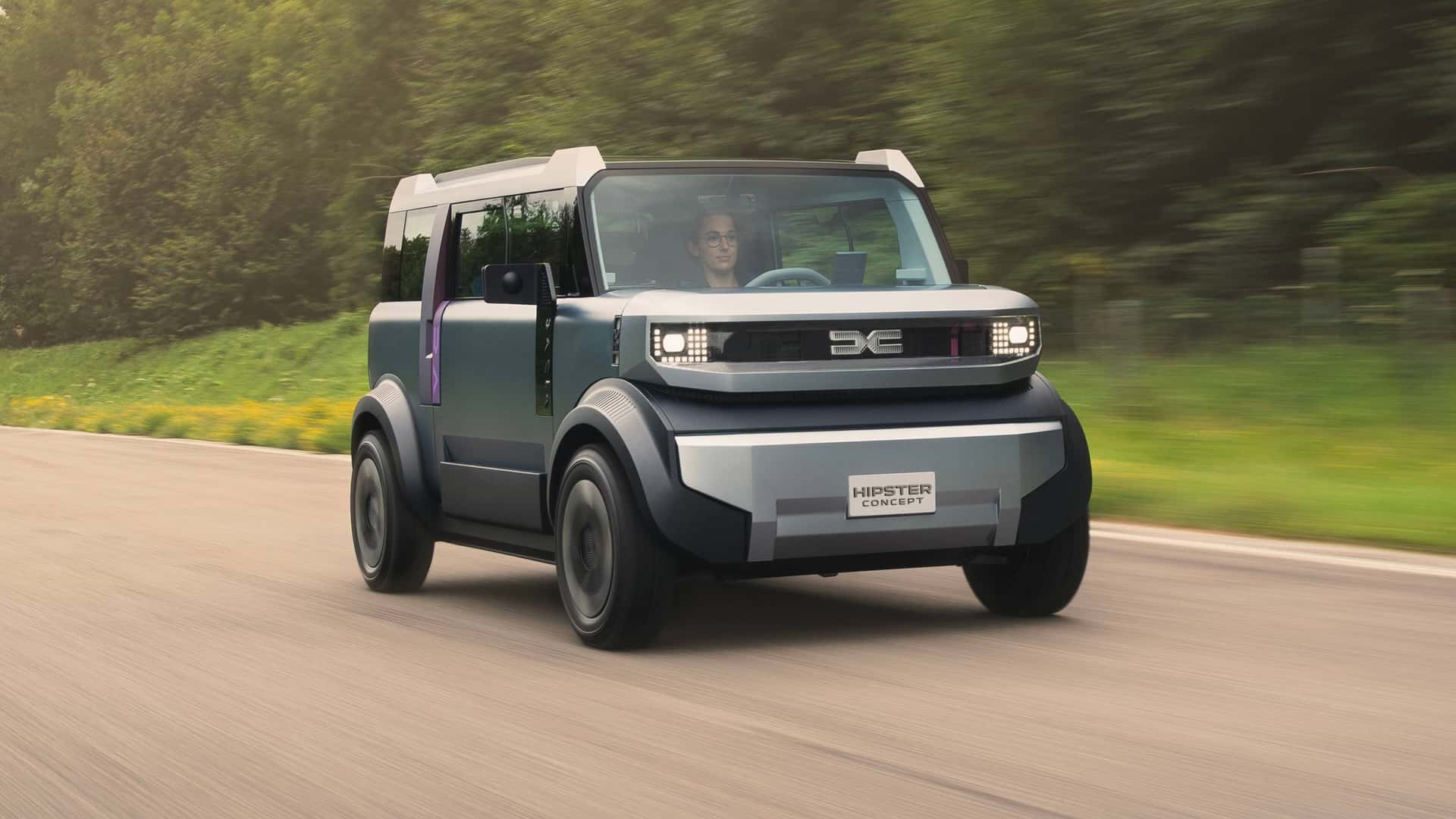 Futuristisk elektrisk bilkoncept kører på en naturskøn vej, med et moderne, kvadratisk design og innovative lysdetaljer.