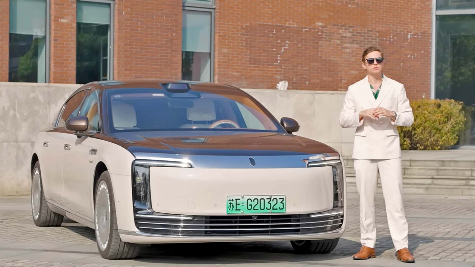 Mand i hvidt jakkesæt ved siden af en luksuriøs elbil foran en murstensbygning