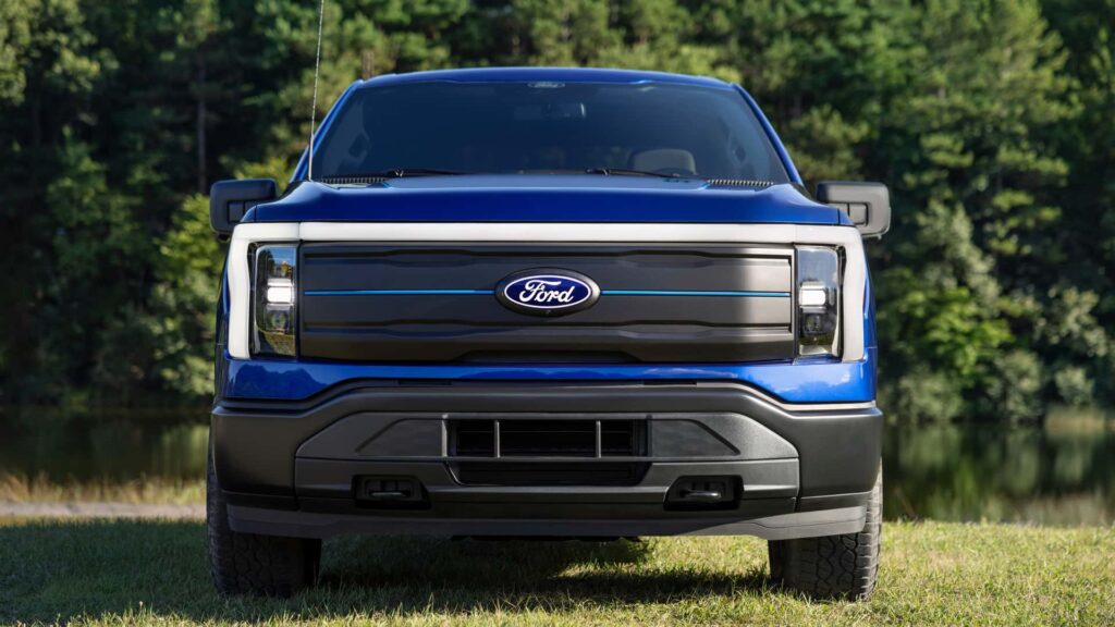 Nærbillede af en blå Ford elbil forfra, parkeret på græs med skov i baggrunden