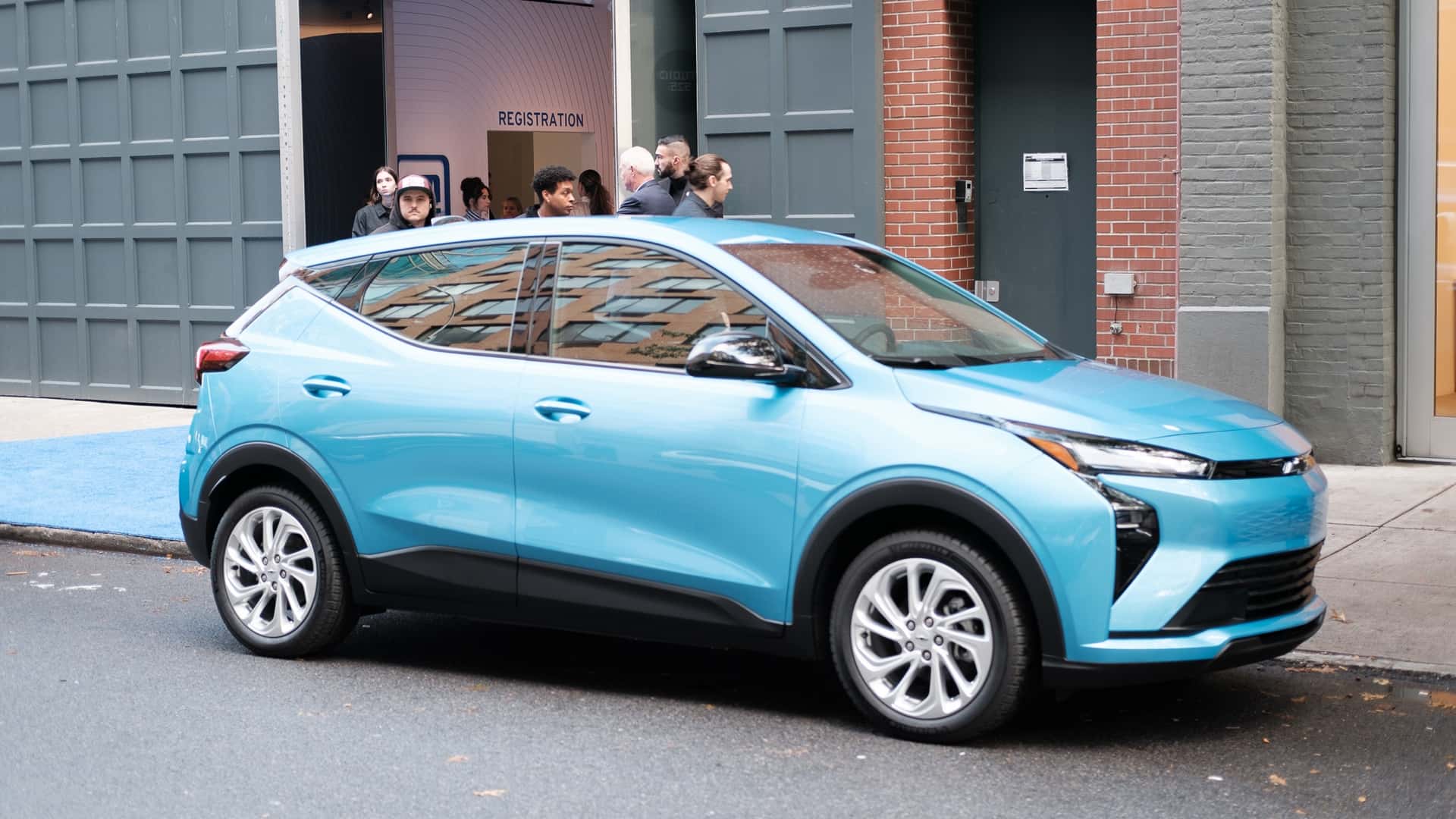 Lyseblå elektrisk bil parkeret foran en bygning med folk udenfor