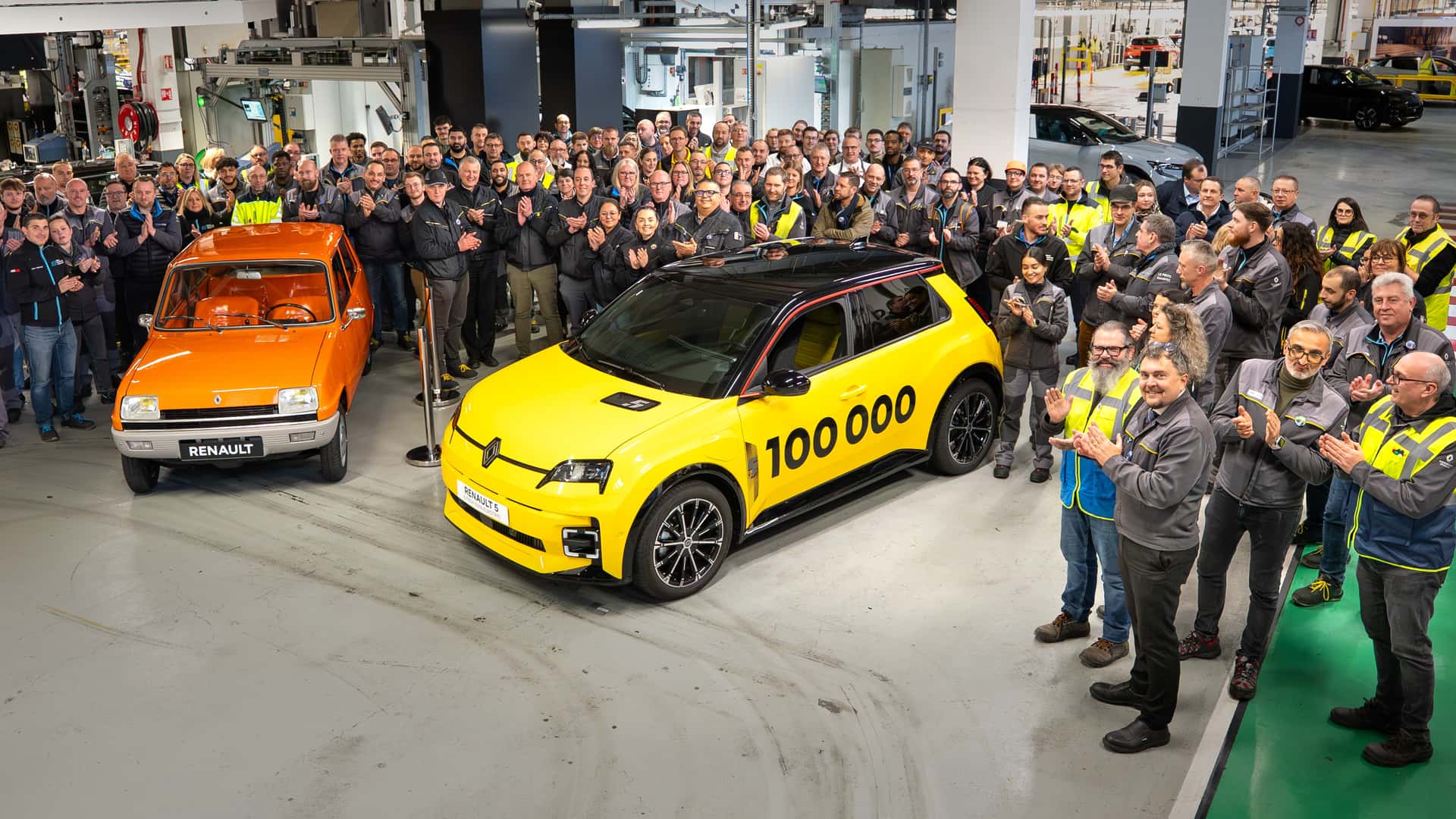 Mennesker samlet omkring gule og orange Renault biler i en fabrikshal, fejrer produktion af 100.000 køretøjer
