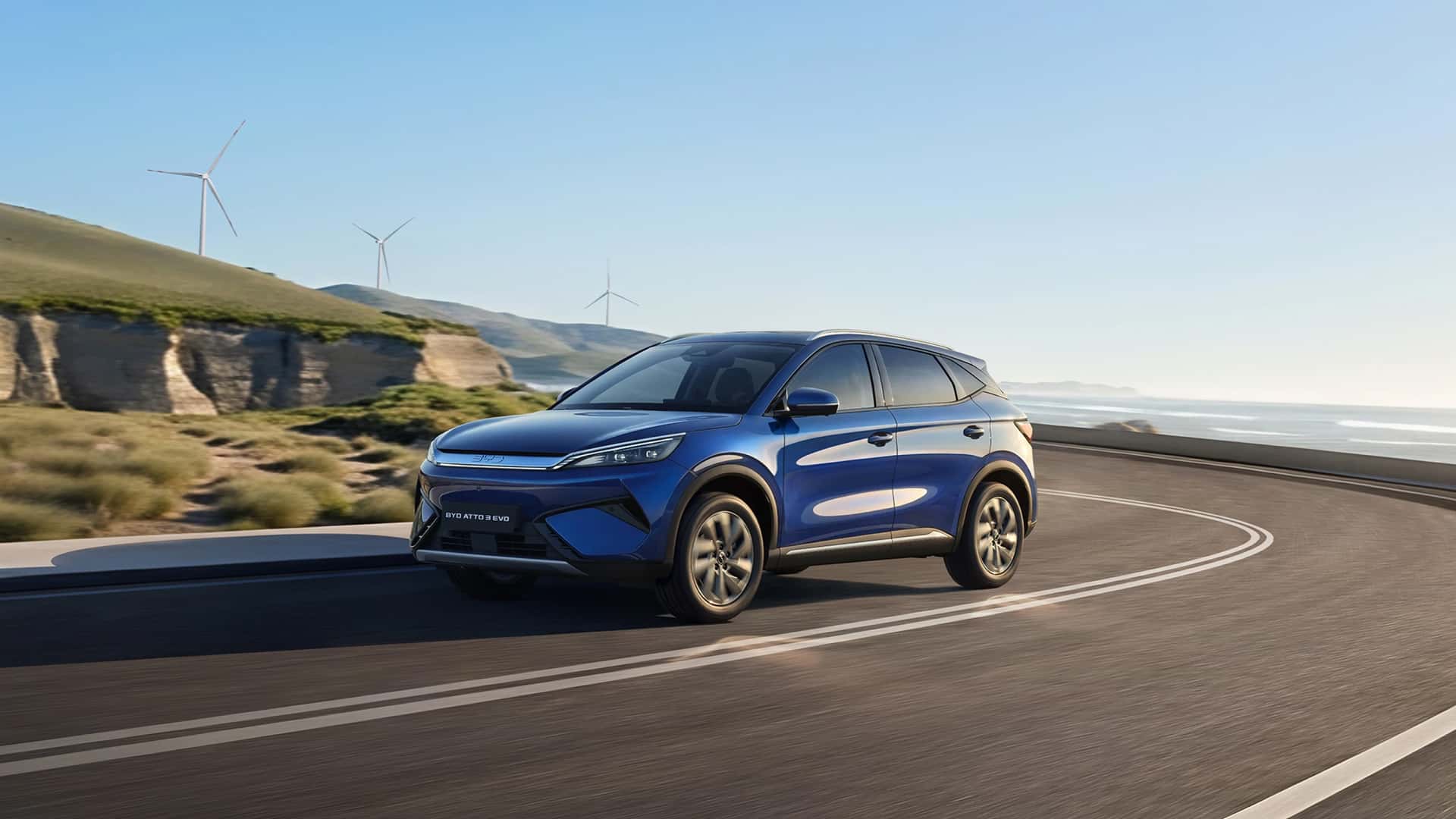 Blå elbil kører på en naturskøn vej med vindmøller i baggrunden