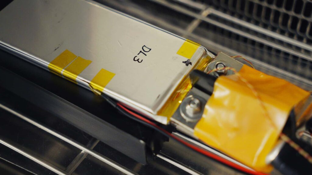 Nærbillede af en Tesla battericelle mærket med 'D13' og omviklet med gul tape, set i et teknisk laboratorium.