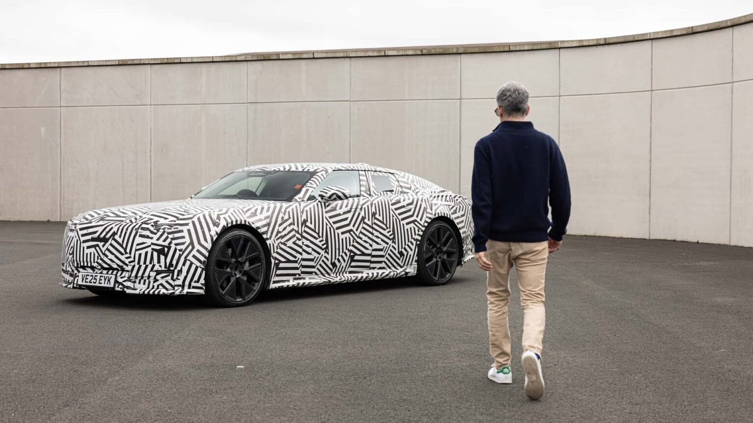 Mand går mod en camouflage-mønstret prototype af en elektrisk bil parkeret på en åben plads.