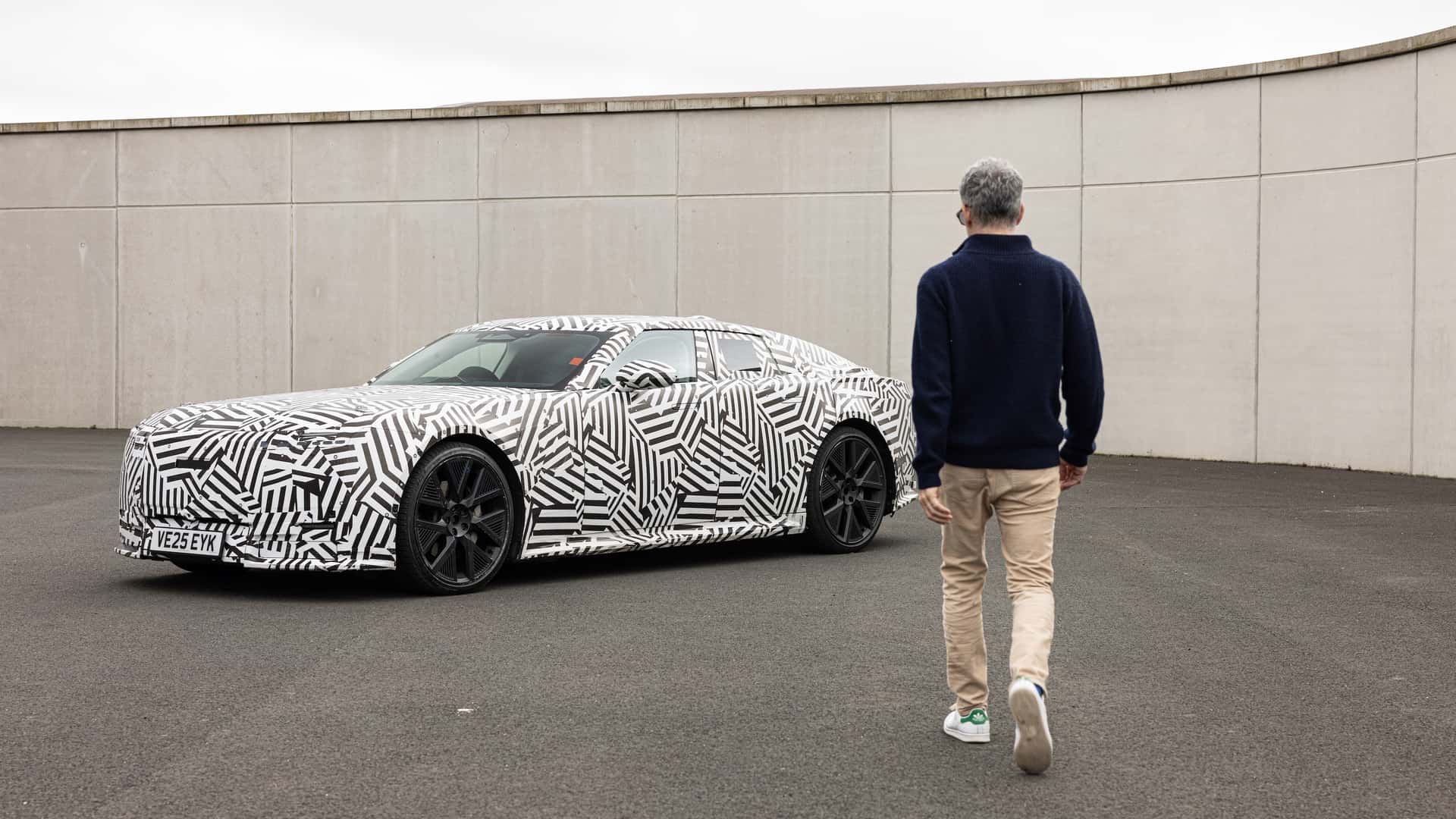 Mand går mod en camouflage-mønstret prototype af en elektrisk bil parkeret på en åben plads.
