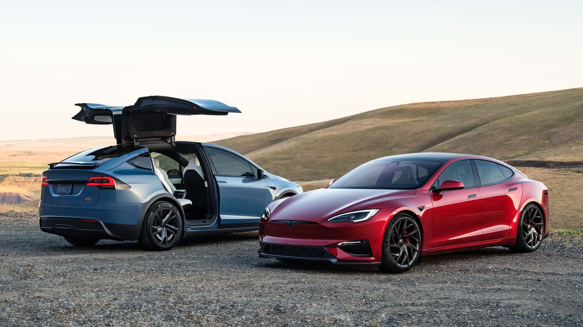 To Tesla-biler parkeret i naturskønt landskab, en blå Model X med døre åbne og en rød Model S