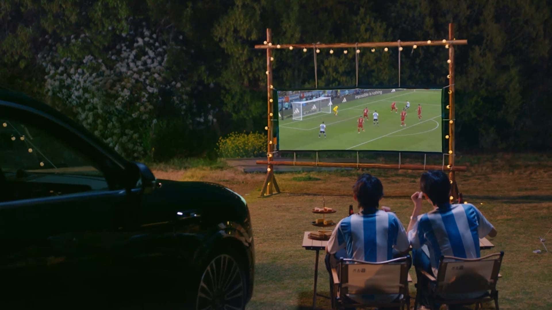 To personer ser fodbold på en udendørs skærm ved siden af en bil