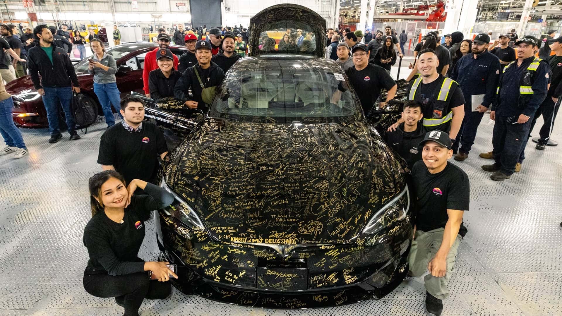 Tesla-bil dækket med underskrifter omringet af smilende medarbejdere i fabriksomgivelser