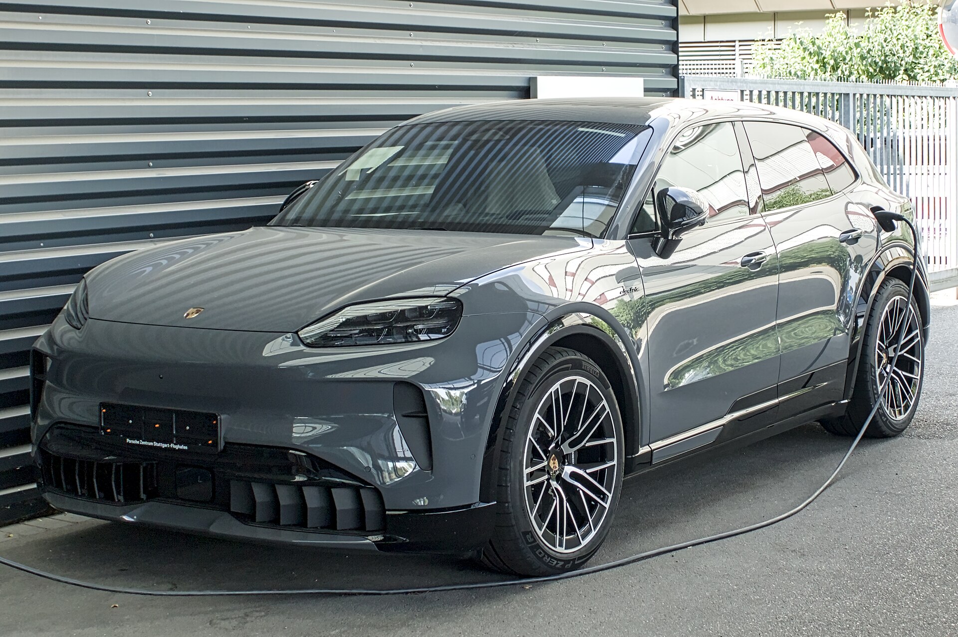 Grå Porsche Cayenne Electric parkeret foran en metalvæg med ladekablet sat i