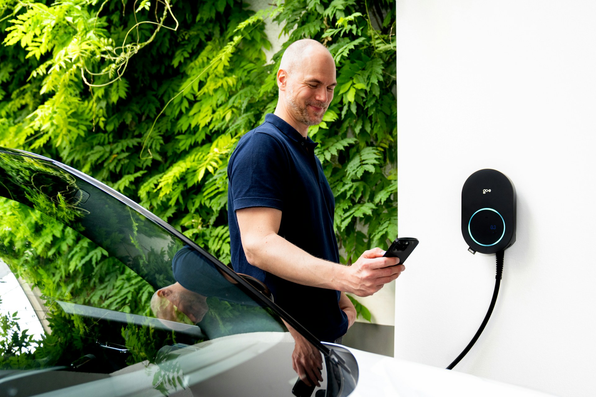 Mand med mobiltelefon står ved en elbil og en ladeboks på husmuren